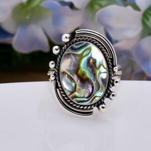 May include: A silver ring with a large oval abalone shell inlay. The shell has iridescent green, blue, and purple colors. The ring is set with small silver beads around the edge of the shell.