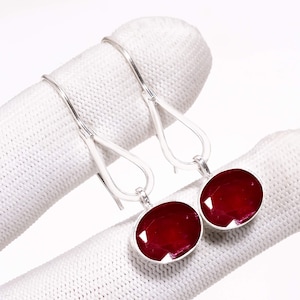 Puede incluir: Un par de pendientes colgantes de plata con piedras preciosas ovaladas de color rojo intenso. Las piedras están engastadas en un bisel de plata, presentadas sobre un fondo blanco.