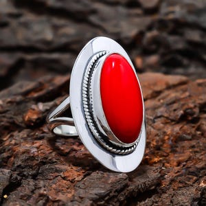 Puede incluir: Un anillo de plata con una piedra grande, ovalada y de color rojo brillante. La piedra está engastada en un bisel de plata pulida con un borde en forma de cuerda. El anillo tiene un diseño dividido.