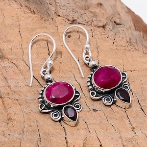 May include: A pair of silver drop earrings with magenta and dark red gemstones. Each earring has an oval magenta stone and a smaller, pear-shaped, dark red stone. The earrings have decorative silver scrollwork around the gemstones.