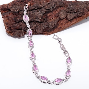 Pode incluir: Uma pulseira de prata com pedras rosa ovais e pequenos detalhes transparentes. A pulseira &eacute; exibida sobre um fundo texturizado marrom escuro, destacando seu design e brilho. O fecho &eacute; vis&iacute;vel, indicando a usabilidade da pulseira.