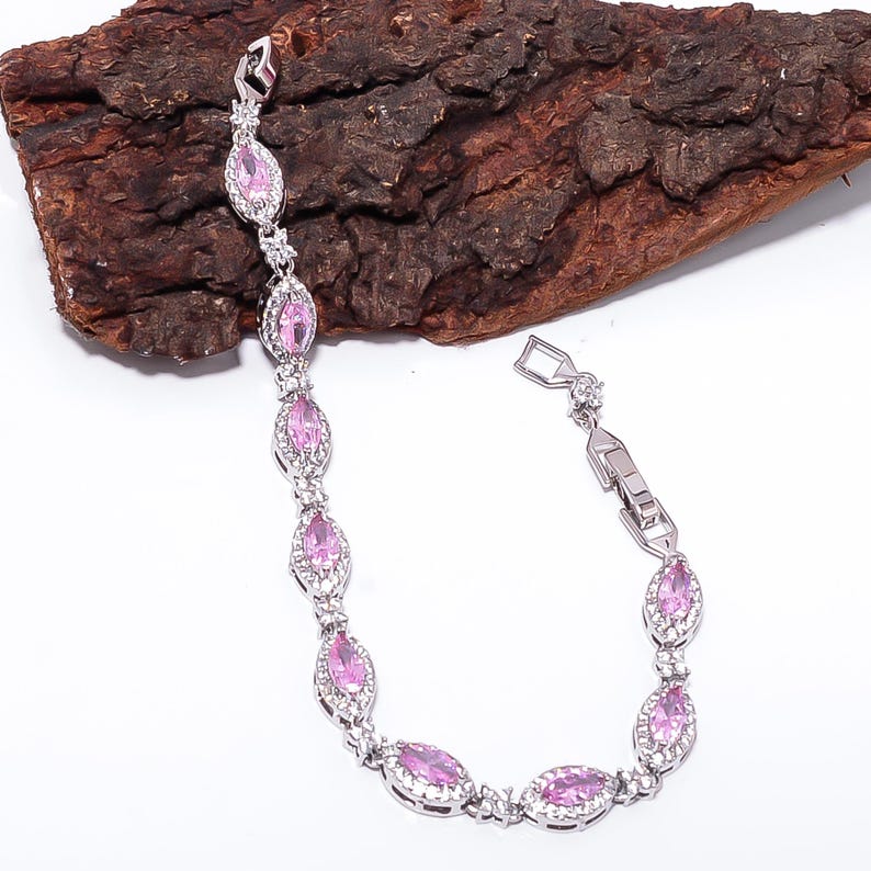 Pode incluir: Uma pulseira de prata com pedras rosa ovais e pequenos detalhes transparentes. A pulseira &eacute; exibida sobre um fundo texturizado marrom escuro, destacando seu design e brilho. O fecho &eacute; vis&iacute;vel, indicando a usabilidade da pulseira.