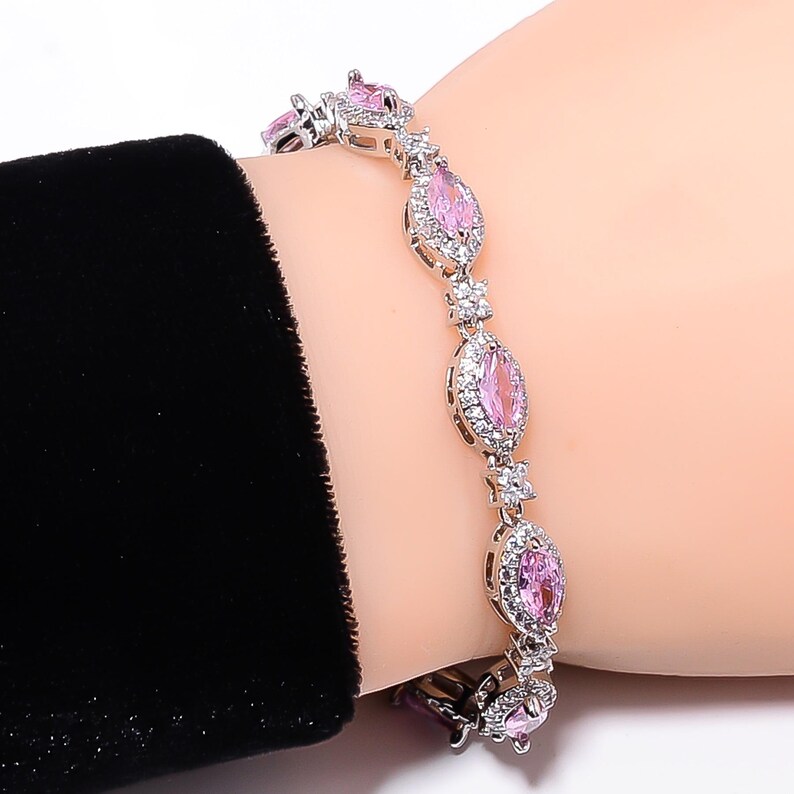 Pode incluir: Uma pulseira de prata com pedras preciosas rosa em forma oval e pequenas pedras brancas. A pulseira est&aacute; em um pulso, com uma manga preta vis&iacute;vel ao fundo. As pedras s&atilde;o cravejadas em metal prateado.