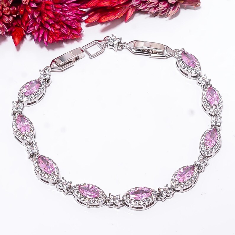Pode incluir: Uma pulseira de prata com pedras preciosas rosa ovais, cada uma rodeada por pedras claras menores. A pulseira est&aacute; sobre um fundo branco, com flores rosa no canto superior esquerdo. A pulseira tem um fecho seguro. As pedras medem cerca de 1 cm.