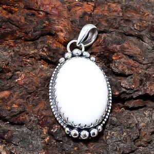 Puede incluir: Colgante ovalado blanco con marco de metal plateado. El colgante tiene un centro de piedra blanca lisa y pulida, rodeado por un borde detallado y texturizado. La argolla es un dise&ntilde;o de bucle simple. El colgante se muestra sobre una superficie marr&oacute;n texturizada.