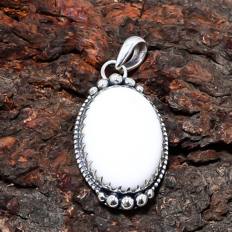 Puede incluir: Colgante ovalado blanco con marco de metal plateado. El colgante tiene un centro de piedra blanca lisa y pulida, rodeado por un borde detallado y texturizado. La argolla es un dise&ntilde;o de bucle simple. El colgante se muestra sobre una superficie marr&oacute;n texturizada.