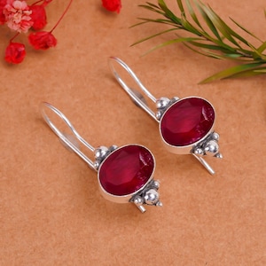 Può includere: Un paio di orecchini pendenti in argento con pietre preziose rosse di forma ovale. Gli orecchini hanno un design semplice con piccole perle d'argento intorno alle pietre preziose.