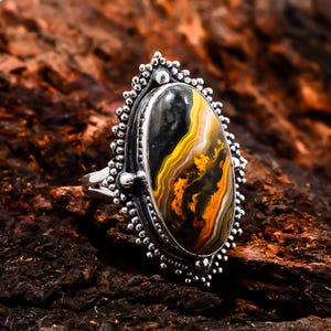 May include: Silver ring with an oval-shaped gemstone. The gemstone features a pattern of black, yellow, and orange colors. The ring has a detailed, beaded border and a simple band.