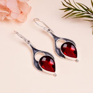 May include: A pair of silver dangle earrings with red gemstone drops. The earrings are teardrop shaped and have a delicate design.