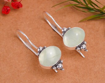 May include: A pair of silver earrings with oval, pale green gemstones. The earrings have a hook design and are adorned with small, round silver accents. The gemstones have a smooth, polished appearance. The earrings are displayed on a brown surface.