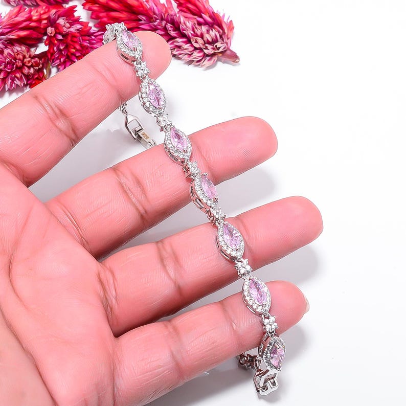 Pode incluir: Uma pulseira de prata com pedras preciosas ovais rosa claro e pequenas pedras de acento transparentes. A pulseira &eacute; segurada em uma m&atilde;o, com flores rosas ao fundo. &Eacute; uma joia.