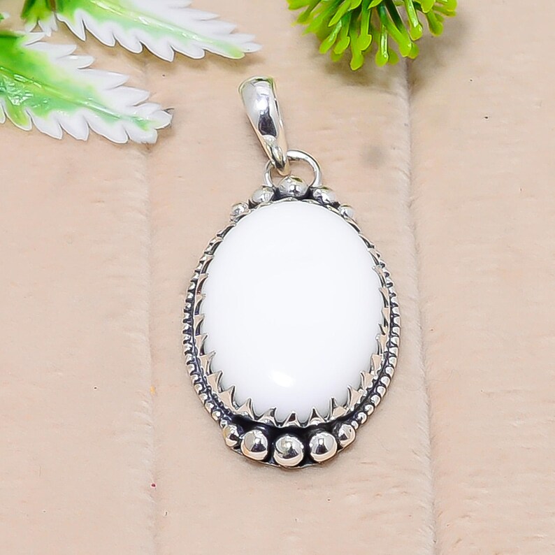 Puede incluir: Colgante ovalado blanco con una argolla plateada y borde decorativo. El colgante tiene una superficie lisa y pulida y un marco detallado y texturizado. La argolla es un simple lazo para sujetar a una cadena.