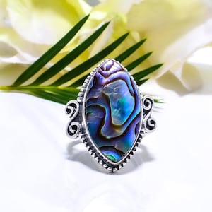 Puede incluir: Un anillo de plata con una gran concha de abulón iridiscente engastada en forma de marquesa. El anillo tiene un diseño detallado y ornamentado alrededor de la concha.