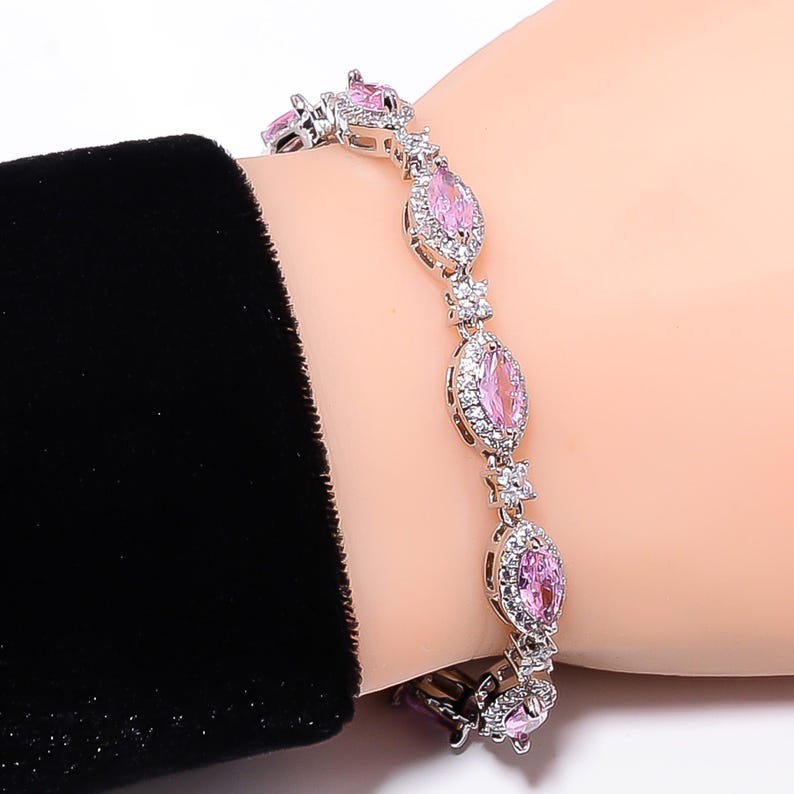 Pode incluir: Uma pulseira de prata com pedras preciosas rosa em forma oval e pequenas pedras brancas. A pulseira est&aacute; em um pulso, com uma manga preta vis&iacute;vel ao fundo. As pedras s&atilde;o cravejadas em metal prateado.