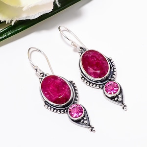 May include: A pair of silver dangle earrings featuring oval-cut, deep pink gemstones. Each earring has a larger oval stone at the top and a smaller round stone below, set in a decorative silver frame. The earrings are displayed against a white background.