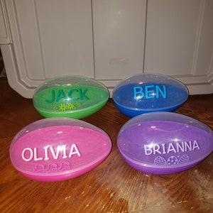 May include: Four clear plastic Easter eggs with different colored bases and names written on them. The names are Jack, Ben, Olivia, and Brianna.