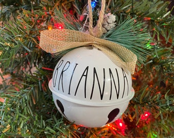 May include: White ceramic Christmas ornament shaped like a bell with the name "Brianna" written in black script. The ornament is decorated with black polka dots and has a brown burlap bow and pine sprigs.