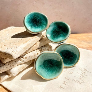 May include: Four teal ceramic knobs with a crackle glaze and gold trim, arranged on a stack of light-coloured stones. The knobs are on a white cloth with the text "THE GARDEN Workshop".