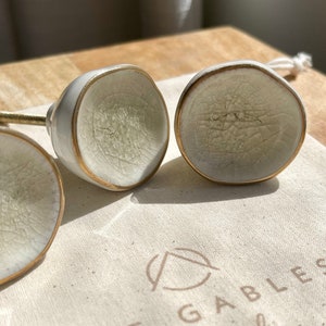 May include: Four white ceramic cabinet knobs with gold edging and a crackle glaze pattern are arranged on a canvas bag. The bag has the text "The Gables Workshop" printed on it. The knobs are on a wooden surface.