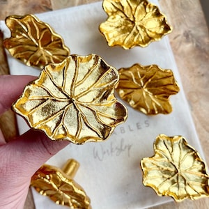 May include: Six gold leaf-shaped cabinet knobs with a textured surface. The knobs are arranged on a white fabric surface with the text "Wribbles Workshop" visible.