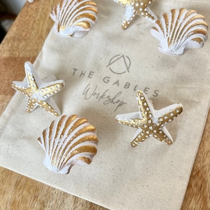 May include: A collection of six decorative items with a coastal theme, featuring starfish and seashell designs in white and gold. They are presented on a beige drawstring bag with the text "THE GABLES Workshop". The background is a wooden surface.
