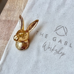 May include: A gold-coloured rabbit head door knob with a textured surface. The knob is sitting on a white canvas bag with the text "The Gables Workshop" printed on it.