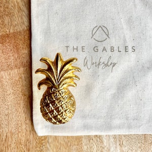 May include: A gold pineapple-shaped door knocker with a textured surface. The knocker is on a white canvas bag with the text "THE GABLES Workshop" printed on it.