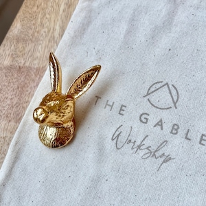 May include: A gold-coloured rabbit head door knob with a textured surface. The knob is sitting on a white canvas bag with the text "The Gables Workshop" printed on it.