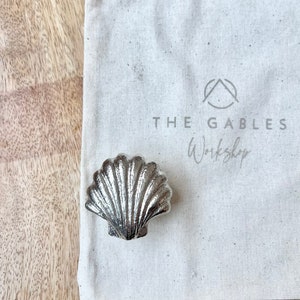 May include: A silver seashell ring holder with a textured surface. The ring holder is sitting on a white canvas bag with the text "THE GABLES Workshop" printed on it.
