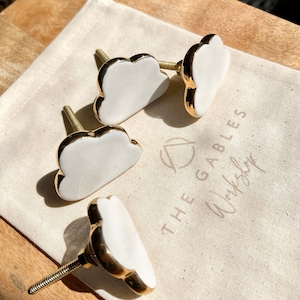 May include: Four white cloud-shaped cabinet knobs with gold trim and threaded posts, displayed on a canvas bag. The bag has the text "THE GABLES Workshop" printed on it. The knobs are on a wooden surface.