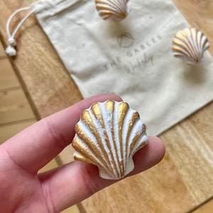 May include: Three white seashells with gold accents. The seashells are on a white fabric bag with the text "The Gables Workshop" printed on it.