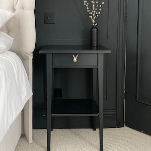 May include: Black bedside table with a single drawer and a lower shelf. The drawer has a silver rabbit knob. The table is in a room with a white headboard and a vase of white flowers.