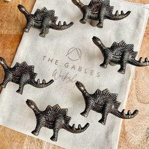 May include: Five antique bronze dinosaur-shaped cabinet knobs with a textured finish. The knobs are arranged on a white canvas bag with the text "THE GABLES Workshop" printed on it.