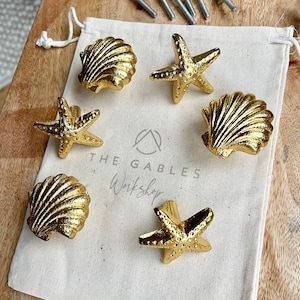 May include: Six gold cabinet knobs in seashell and starfish designs. The knobs are on a white drawstring bag with the text "THE GABLES Workshop". Mounting hardware is visible above the bag.