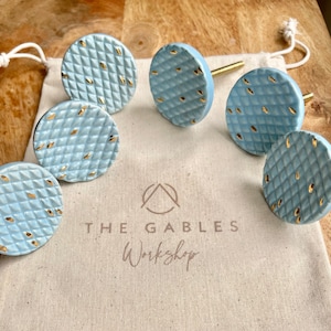 May include: Set of six light blue ceramic cabinet knobs with gold flecks. The knobs are in a white drawstring bag with the text "THE GABLES Workshop" printed on it.