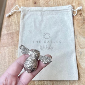 May include: A silver bee-shaped door knob with a textured surface. The knob is held in a hand and is in front of a white canvas bag with the text "THE GABLES Workshop" printed on it.