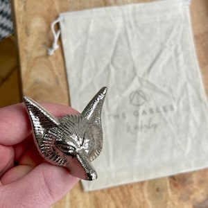 May include: Silver fox head bottle stopper with a detailed design. The stopper is in a small white drawstring bag with the text "The Gables Distillery" printed on it.