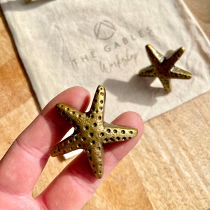 May include: Four gold-toned starfish drawer pulls with a textured surface. The starfish are on a white fabric surface with the text "THE GABLES Workshop" printed on it.