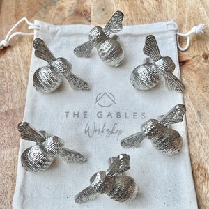 May include: Five silver bunny-shaped decorative objects with textured bodies and wing-like ears are displayed on a white drawstring bag. The bag has the text "THE GABLES Workshop" printed on it. The objects are on a light brown wooden surface.