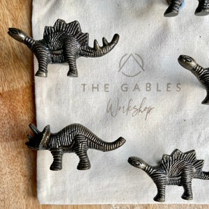 May include: Four metal dinosaur-shaped drawer pulls in a rustic bronze finish. The pulls are displayed on a white fabric surface with the text "The Gables Workshop".