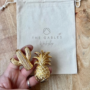 May include: Gold-coloured cactus and pineapple shaped decorative objects. The objects are held in front of a canvas bag with the text "THE GABLES Workshop". The objects are highly detailed and have a textured surface.