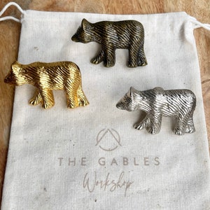 May include: Three bear-shaped drawer pulls in gold, silver, and bronze finishes. The pulls are displayed on a white fabric surface with the text "The Gables Workshop".