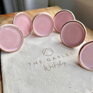 May include: A set of pink ceramic cabinet knobs with gold edging. The knobs are displayed on a wooden surface and a canvas bag with the text "THE GABLES Workshop". The knobs are circular in shape.