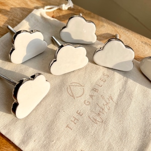 May include: Six white ceramic cloud-shaped drawer pulls with silver metal accents. The pulls are arranged on a white fabric surface with the text "THE GABLES" printed on it.