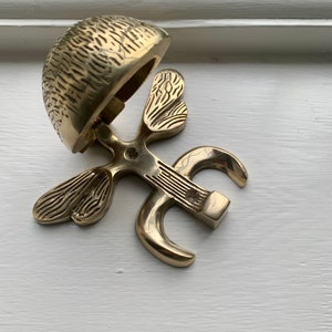 May include: A gold-toned metal door knocker shaped like a stylized butterfly with a bell on top. The bell is open and has a textured surface.