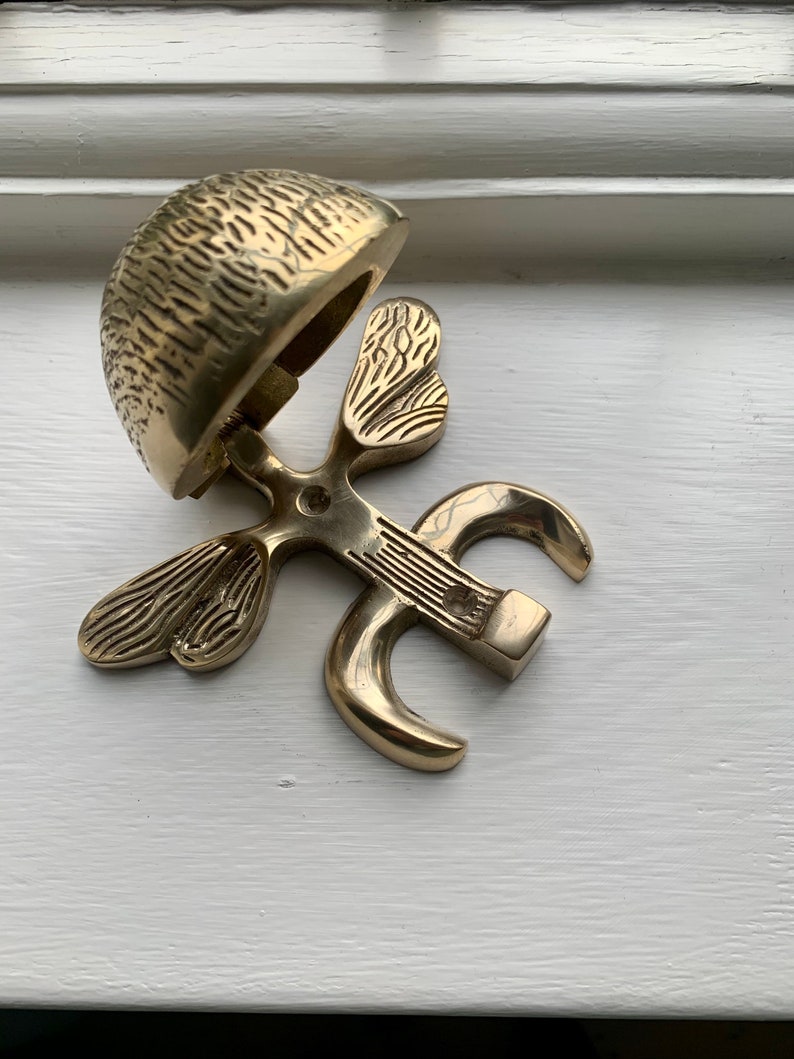 May include: A gold-toned metal door knocker shaped like a stylized butterfly with a bell on top. The bell is open and has a textured surface.