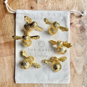 May include: Six gold bee-shaped drawer pulls in a white fabric bag. The bag has the text "THE GABLES Workshop" printed on it.