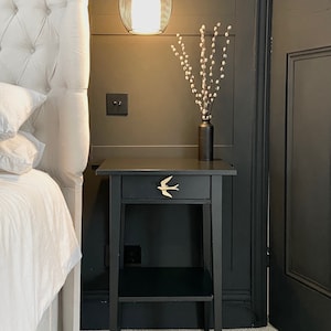 May include: Black bedside table with a single drawer and a lower shelf. The drawer has a gold bird-shaped pull. The table is next to a white headboard and a white bed.