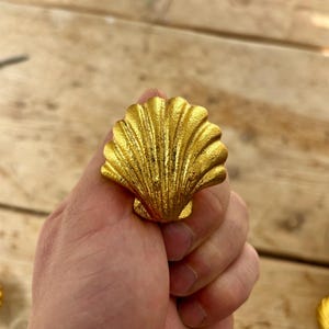 May include: A gold-coloured seashell-shaped decorative object. The seashell has a textured surface and is held in a hand. The background is a wooden surface.