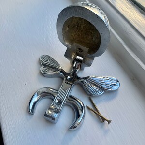 May include: A silver door knocker shaped like a butterfly with two wings. The knocker is open and shows the inside of the knocker, which is hollow. Two screws are included.